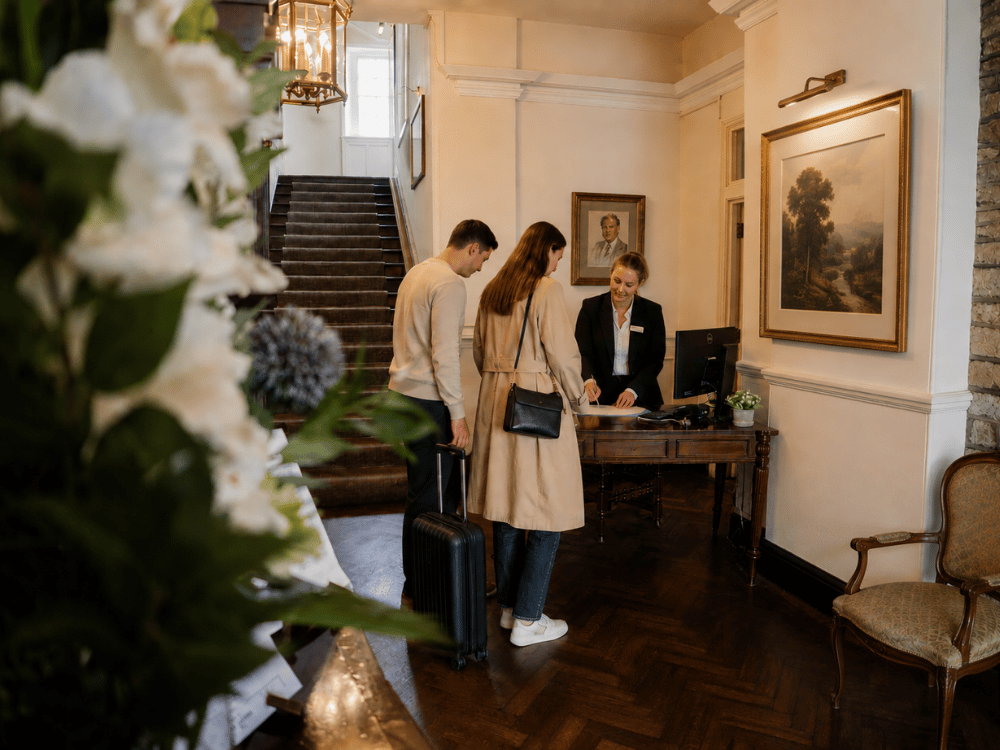 Hotel lobby check-in: two guests with luggage at a wooden desk speaking with a receptionist under warm lighting.